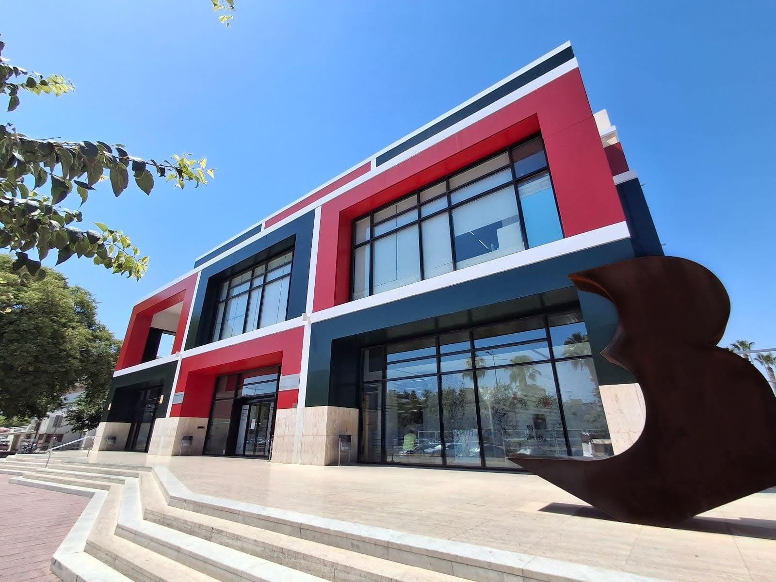 Edificio contemporáneo de dos plantas con fachada roja, azul y blanca, grandes ventanales, escaleras de acceso y una escultura abstracta metálica en el exterior bajo un cielo despejado. Es la sede de la Biblioteca Regional de Murcia.