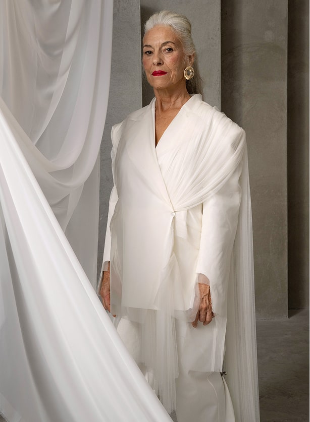 Mujer mayor vestida de blanco, de pie junto a una cortina.